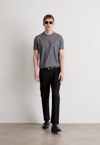 Γκρι κοντομάνικο t-shirt συνδυασμένο με μαύρα παντελόνια και μαύρα παπούτσια. Μοντέλο που φοράει γυαλιά ηλίου, στέκεται μπροστά από λευκό φόντο.