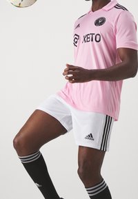 Vaaleanpunainen jalkapallopaita mustalla logolla ja tekstillä, valkoiset shortsit mustilla raidoilla, mustat polveen ulottuvat sukat valkoisilla raidalla.