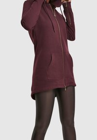 Femme portant un long sweat à capuche bordeaux zippé avec poches avant et manches avec trous pour les pouces, associé à des collants noirs transparents sur un fond uni.