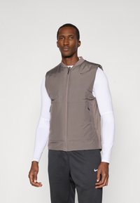 Gilet marrone senza maniche con zip frontale, tasche laterali e colletto alto. Realizzato in tessuto leggero, abbinato a una camicia bianca a maniche lunghe.