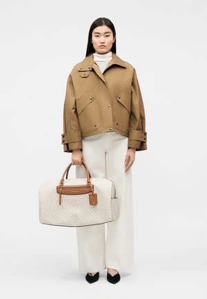 Γυναίκα που φοράει ένα μεγάλο καφέ μπουφάν και λευκό παντελόνι, κρατώντας μια μπεζ τσάντα ταξιδίου Michael Kors με καφέ χερούλια και λεπτομέρειες.