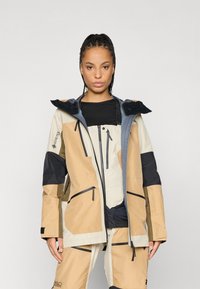 GORE-TEX-Jacke in Beige, Braun und Marineblau mit Kapuze, Reißverschlüssen und mehreren Taschen; elegantes Design und strukturierte Stoffoberfläche.