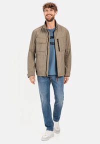 Beige leichte Jacke mit mehreren Taschen, einer mit Reißverschluss versehenen Brusttasche, kombiniert mit einem blauen T-Shirt und blauen Jeans, getragen mit weißen Sneakers.