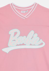 Rozā sieta krekls ar baltām un gaiši ziliem svītrainiem V veida kakla izgriezumiem. Iespēts "Barbie" lielā baltā burtnīcā un zem teksta ir izliekta balta akcenta līnija.