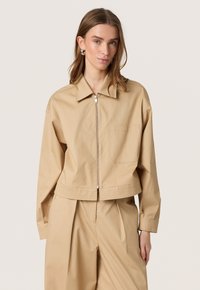 Lys beige cropped jakke lavet af bomuld med spidse kraver, frontlynlås, en brystlomme og let løse ærmer.
