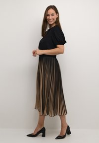 Juoda ir smėlio spalvos klostuota suknelė su trumpomis pūstomis rankovėmis, paryškinta juosmeniu, su plačiu sijonu, derinama su juodomis aukštakulnėmis bateliais.