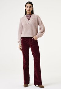 Helles rosa geripptes Strickpullover mit V-Ausschnitt, über einem floralem Oberteil getragen, kombiniert mit burgunderfarbenen Cordflared-Hosen und hellbraunen Absatzschuhen.