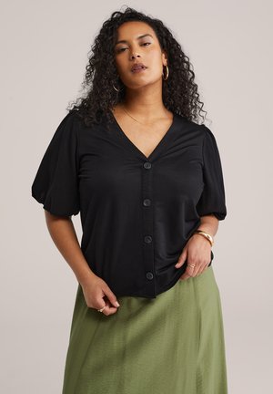 Frau mit lockigem Haar, die goldene Creolen, eine schwarze Bluse mit Puffärmeln und einen olivgrünen Rock trägt, vor einem schlichten Hintergrund stehend.