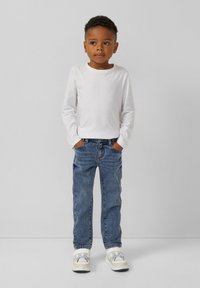 Camicia bianca a maniche lunghe, jeans blu in denim e sneakers bianche. I jeans hanno una vestibilità classica con una texture sbiadita e tasche standard.