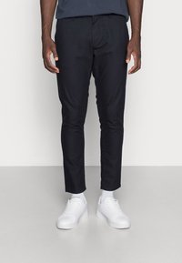Navy slim-fit chinos made of cotton blend fabric, featuring a flat front design, side pockets, and styled with white sneakers and ankle socks.