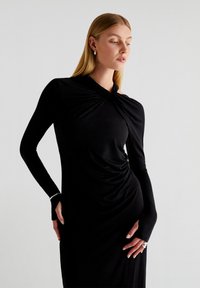 Robe noire longue avec des manches longues et un décolleté torsadé. Tissu lisse et extensible avec une silhouette ajustée et des accents de trou pour le pouce.