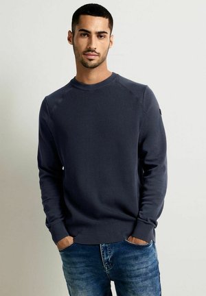 Marineblaue, gerippte Strickjacke mit langen Ärmeln, Rundhalsausschnitt und lockerer Passform, kombiniert mit blauen Jeans. Dezentes Logo-Detail am Ärmel.
