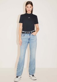 Camiseta negra de manga corta con logo, jeans rectos de color azul claro, cinturón negro y zapatillas blancas con detalles en negro. Textura suave.