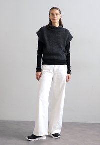 Pull noir texturé à col montant superposé sur un haut noir à manches longues, associé à un pantalon large blanc et des baskets noires.