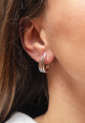 Primo piano dell'orecchio di una persona con tre orecchini a cerchio d'argento indossati sul lobo, capelli parzialmente visibili, pelle liscia e chiara.