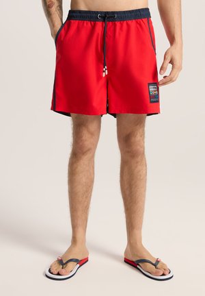Czerwone spodenki kąpielowe z granatowym pasem, sznurkiem ściągającym i bocznymi paskami; z przodu znajduje się naszywka z grafiką. W połączeniu z klapkami.