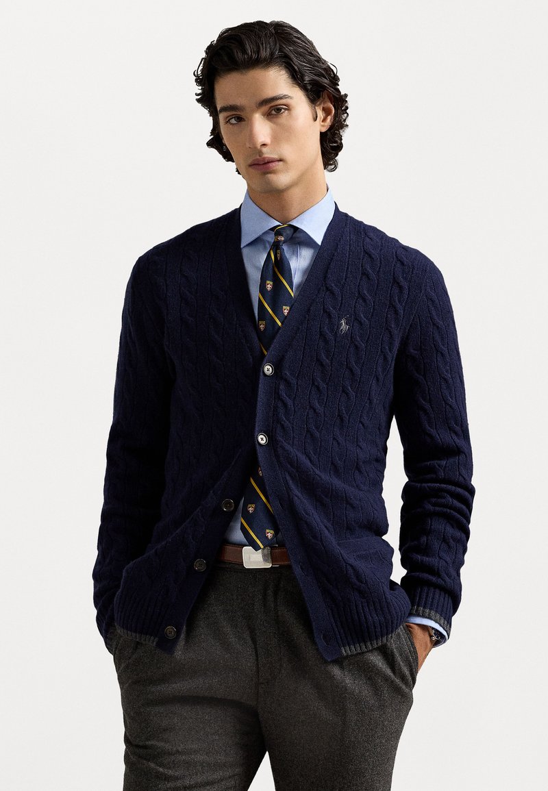 Cardigan en tricot à câbles marine avec cinq boutons, porté sur une chemise bleue et une cravate à motifs. Pantalon gris et ceinture brune, présentant un design épuré.