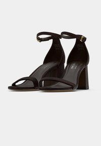 Καφέ σατινέ πέδιλα με τακούνι, λουρί στον αστράγαλο, ανοιχτή μύτη και ορθογώνιο τακούνι. Λείο υλικό και μινιμαλιστικές λεπτομέρειες.