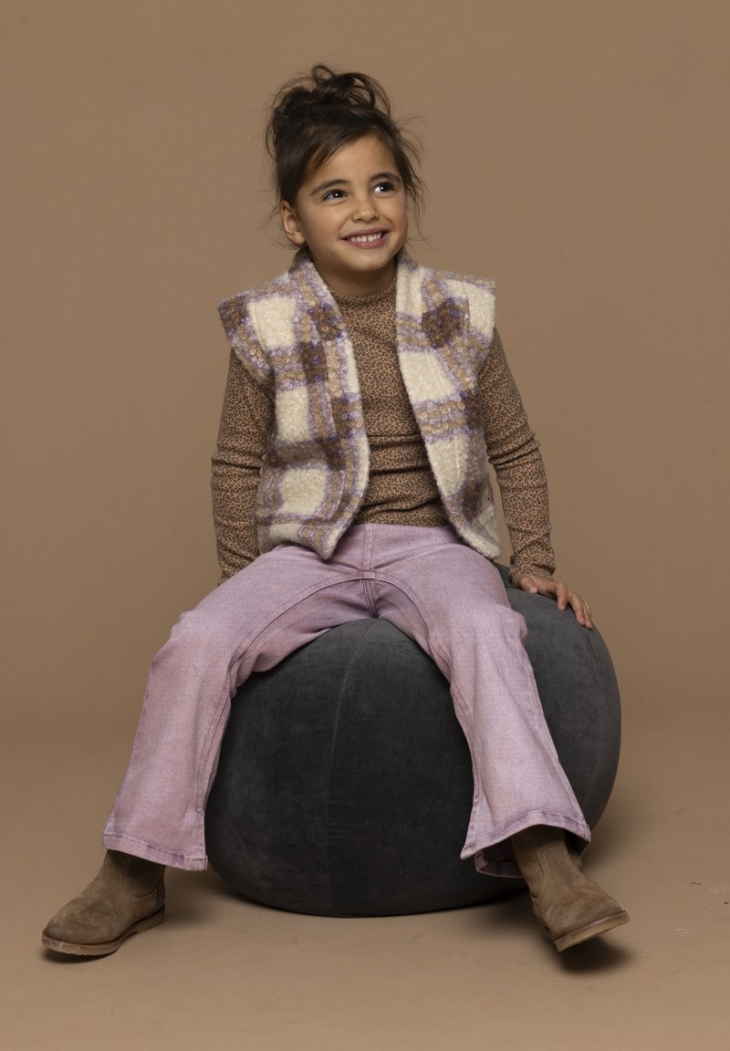 Witte en paarse geruite vest over een bedrukte bruine top, gecombineerd met roze uitlopende broek en tan laarzen, zittend op een donker rond kussen.