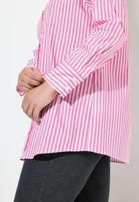 Roze en witte gestreepte langemouwen shirt met een knoopmanchet, gecombineerd met donkergrijze leggings, vanaf de zijkant weergegeven.
