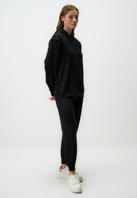 Zwarte hoodie met lange mouwen, ontspannen pasvorm, gecombineerd met bijpassende zwarte joggingsbroek met elastische manchetten en witte sneakers.