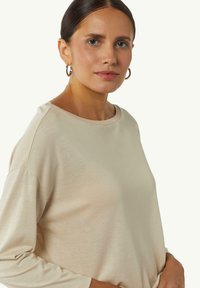 Helles beige Langarmshirt mit rundem Ausschnitt und leicht gesenkten Schultern, aus weichem, strukturiertem Stoff. Einfaches Design ohne Muster.