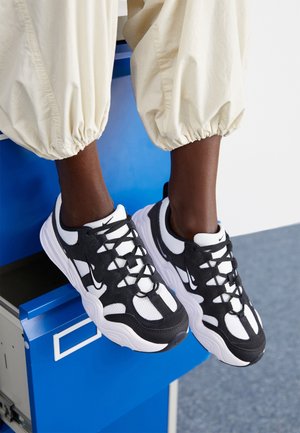Personne portant des baskets Nike noires et blanches avec des lacets noirs, assise sur un casier métallique bleu, pantalon crème resserré aux chevilles.