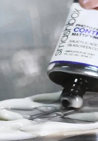 Flacon noir d'un produit Smashbox avec une finition mate, ornée d'une étiquette blanche et d'accents violets, libérant une substance claire semblable à un gel.