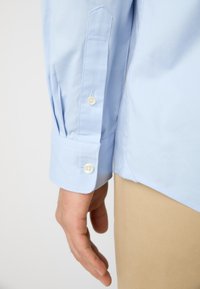 Chemise boutonnière bleu clair à manches longues, avec des boutons blancs et une texture lisse. Détails de poignets visibles et coupe ajustée.
