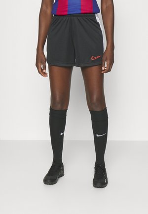 Osoba ubrana w czarne sportowe szorty Nike, czarne podkolanówki z białymi logo, czarne korki oraz niebiesko-czerwony top, stojąca na jednolitym tle.