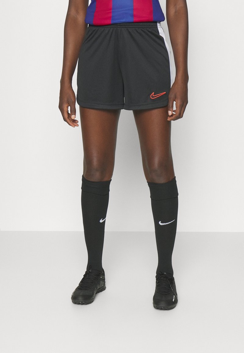Personne portant un short de sport Nike noir, des chaussettes noires montantes avec des logos blancs, des crampons noirs et un haut bleu et rouge, debout devant un fond uni.
