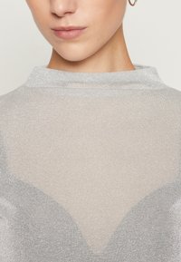 Närbild av en person som bär en genomskinlig silveröverdel och guldörhängen, visar läppar, haka, hals och del av nyckelbenet.