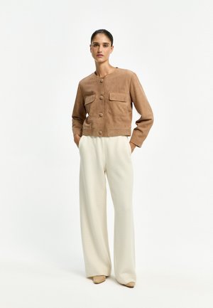 Woman wearing a brown buttoned jacket with flap pockets and loose white wide-leg pants standing with hands in pockets.