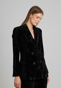 Blazer nero doppiopetto con finitura in velluto testurizzato, dotato di colletto a scialle, tasche frontali e dettagli circolari in metallo argentato.
