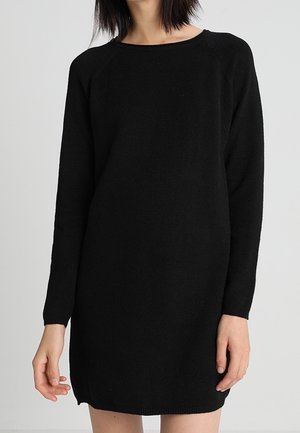 Femme portant une robe noire en tricot à manches longues unie, debout devant un fond gris clair uni.