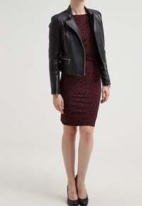 Femme portant une robe mi-longue à motifs rouge et noir, une veste en cuir noir et des chaussures à talons hauts noires, se tenant devant un fond blanc.