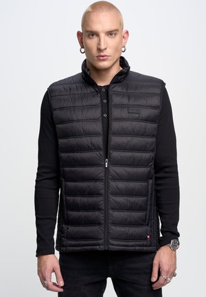 Zwarte puffer vest met horizontale stiksels, hoge kraag en ritssluiting. Bevat een klein logo-embleem en wordt gedragen over een zwart langslijfshemd.