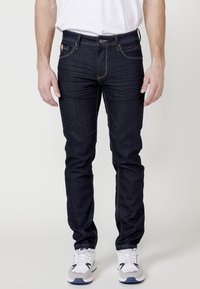 Jean oscuro de mezclilla con un corte ajustado, con costuras amarillas, cinco bolsillos y un cierre de cremallera. Llevados con zapatillas deportivas blancas y grises.