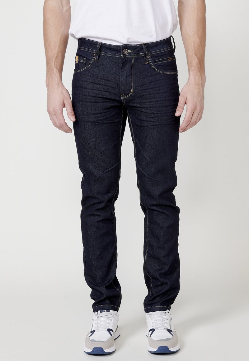 Jean oscuro de mezclilla con un corte ajustado, con costuras amarillas, cinco bolsillos y un cierre de cremallera. Llevados con zapatillas deportivas blancas y grises.