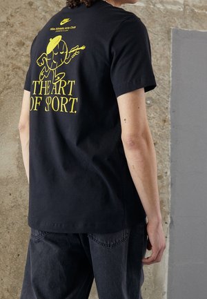 Personne portant un T-shirt noir avec un personnage de basket-ball jaune de style cartoon et le texte « THE ART OF SPORT » au dos, debout contre un mur rugueux.
