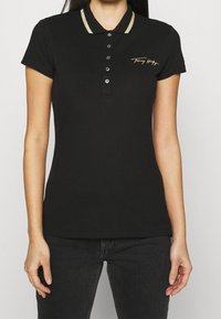 Sort polo-shirt med en guldstribet krave, med en knaplukning og et guldbroderet logo på brystet. Blødt, åndbart bomuldsstof.