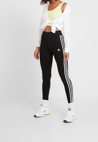 Kvinna som bär svarta Adidas tights med vita ränder, en gul sport-bh, en vit skjorta knuten vid midjan och vita sneakers med blå accenter.