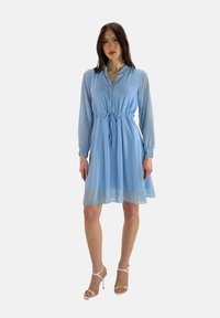 Vestito azzurro chiaro realizzato in tessuto trasparente, con chiusura sul davanti a bottoni, maniche lunghe, vita cintrata con un cordino, lunghezza fino al ginocchio e orlo semi-trasparente.