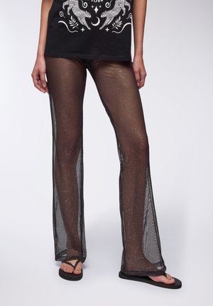Czarne przezroczyste spodnie z błyszczącymi srebrnymi akcentami, szerokimi nogawkami i luźnym krojem. Noszone na czarnej koszulce z grafiką i klapkach.