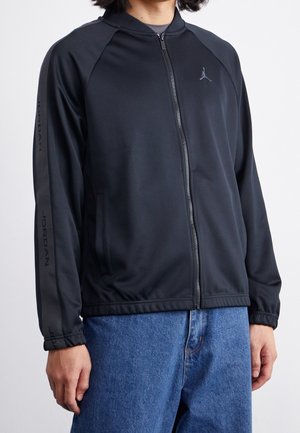Personne portant une veste zippée noire Jordan avec un logo sur la poitrine et le texte « Jordan » sur les manches, associée à un jean bleu.