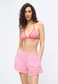 Femeie cu păr lung și creț, purtând un top de bikini roșu cu alb cu model și pantaloni scurți roz deschiși din terry, cu mâinile în buzunare.