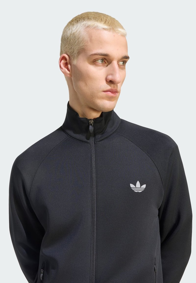 Mann mit kurzen blonden Haaren, der eine schwarze Adidas-Jacke mit Reißverschluss trägt und seitlich vor einem einfarbigen Hintergrund schaut.