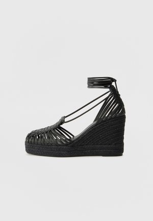 Sandale compensée noire avec brides tressées et fermeture autour de la cheville, dotée d'une semelle plateforme texturée et d'un design à bout ouvert.