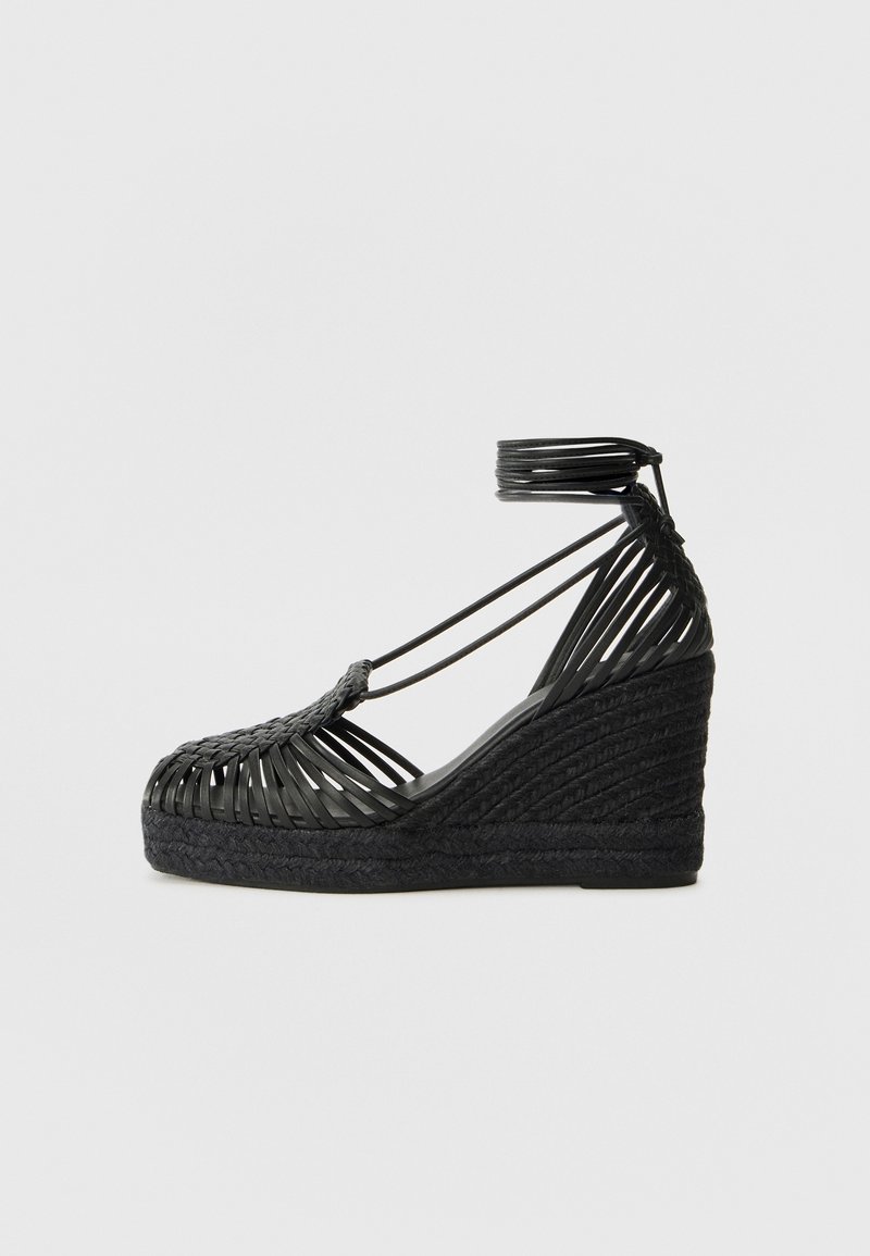 Sandale compensée noire avec brides tressées et fermeture autour de la cheville, dotée d'une semelle plateforme texturée et d'un design à bout ouvert.