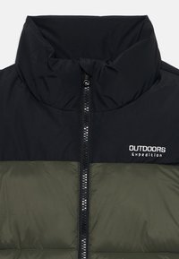 Jakna na napuhavanje s visokim ovratnikom, crnim gornjim dijelom i tamnozelenim donjim dijelom. Ima središnji patentni zatvarač i logo "OUTDOORS Expedition".